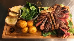 Florentine beef steak on a wooden platter with roast potatoes, bread and fresh salad