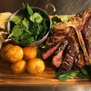 Florentine beef steak on a wooden platter with roast potatoes, bread and fresh salad