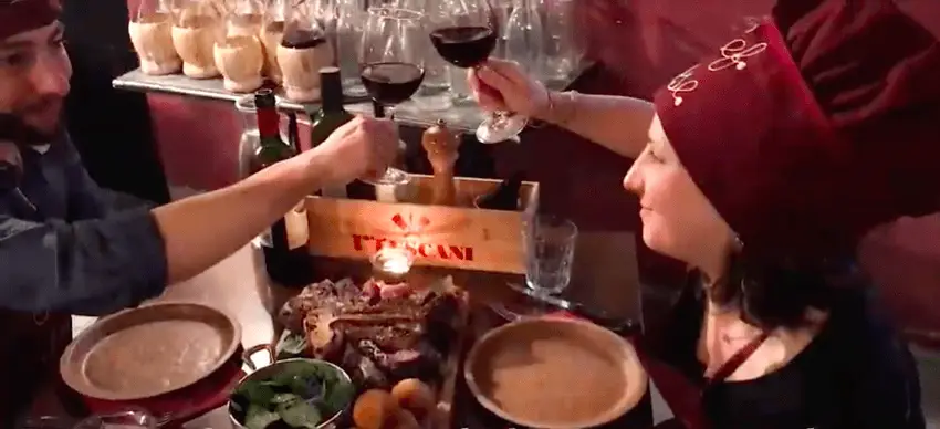 Two wine glasses clink over a platter of Florentine steak and roast potatoes
